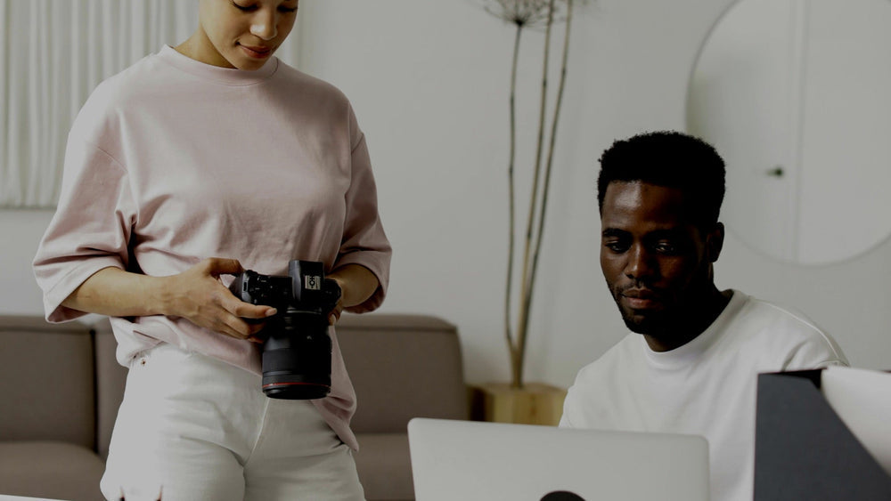 Two creators working together. One standing and holding a camera and the other sitting on a computer.